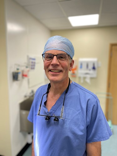 Professor Simon Rogers dressed in blue theatre scrubs with surgical spectacles