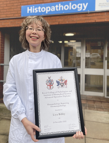 Photo of woman in white labcoat holding a certificate from the Royal College of Pathologists and Institute of Biomedical Science Advanced Specialist Diploma