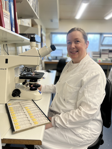 Female in white lab coat sitting at a microscope with a sample tray on the desk