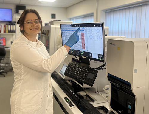 Woman in a white lab coat standing next to a scientific machine pointing at the screen to activate it