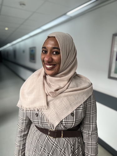 Black woman standing in a hospital corridor wearing a checked brown and cream long sleeved dress with a beige headscarf covering her head and shoulders