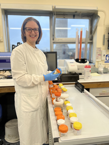 Woman wearing white lab coat holding a small pot with an orange lid standing next to a table full of orange and yellow lidded pots, in a laboratory