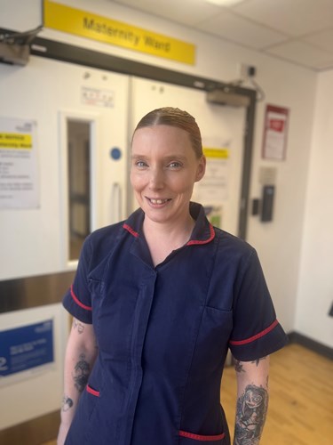 Photo of Rosie Hoyle wearing a ward manager's blue tunic with red piping standing outside the entrance to the maternity ward