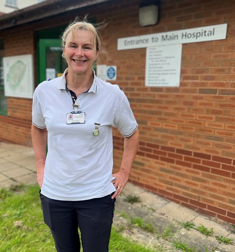 Photo of Helen wearing a white physio polo shirt and navy trouser, blond hair tied up, standing outside entrance to main hospital